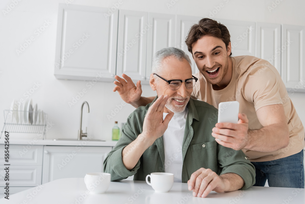 Fototapeta premium cheerful man with adult son waving hands during video call on mobile phone in kitchen