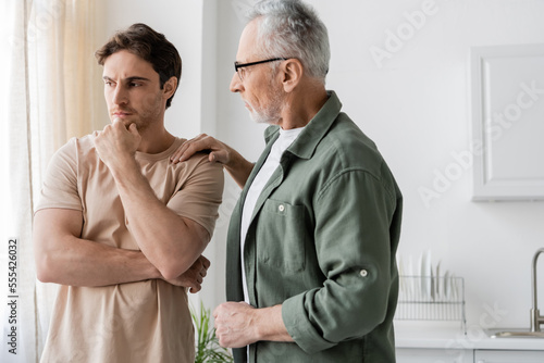 thoughtful and upset man holding hand near face while father calming him in kitchen