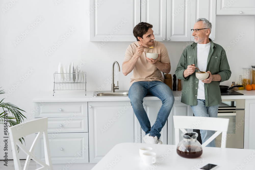 young man sitting on kitchen worktop while having breakfast and talking to smiling dad young man sitting on kitchen worktop while having breakfast and talking to smiling dad