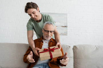 smiling guy holding fathers day present near senior dad sitting on sofa in living room