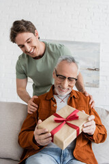 cheerful man hugging shoulders of happy dad holding fathers day present on couch at home