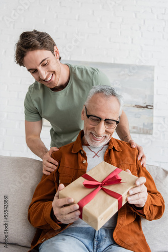cheerful man hugging shoulders of happy dad holding fathers day present on couch at home