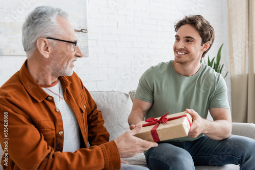 happy man congratulating dad on fathers day and giving him gift box on couch at home