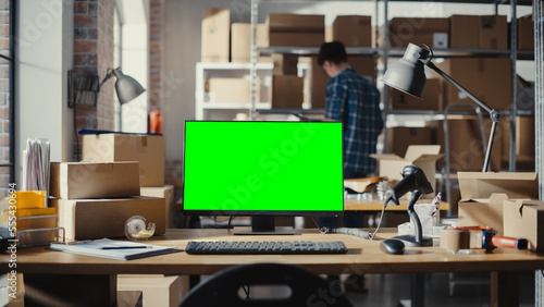 Desktop Computer Monitor Standing on a Table with a Green Screen Chromakey Mock Up Display. Small Business Warehouse with Worker Walking in the Background. Desk with Cardboard Boxes © Gorodenkoff