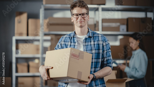 Portrait of a Successful Young Small Business Owner Smiling, Looking at Camera. Caucasian Male Holding a Cardboard Box in a Warehouse Storeroom with Orders Ready for Shipment.