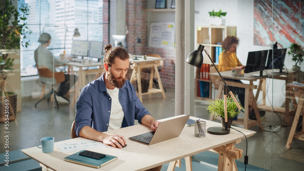 Handsome Long-Haired Bearded Manager Sitting at a Desk in Creative Office. Young Stylish Man Using Laptop Computer in Marketing Agency. Colleagues Working in the Background. High Angle View. Handsome Long-Haired Bearded Manager Sitting at a Desk in Creative Office. Young Stylish Man Using Laptop Computer in Marketing Agency. Colleagues Working in the Background. High Angle View.
