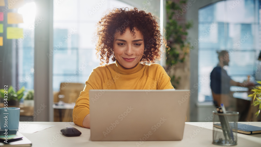 Beautiful Middle Eastern Manager Sitting at a Desk in Creative Office. Young Stylish Female with Curly Hair Using Laptop Computer in Marketing Agency. Colleagues Working in the Background - obrazy, fototapety, plakaty 
