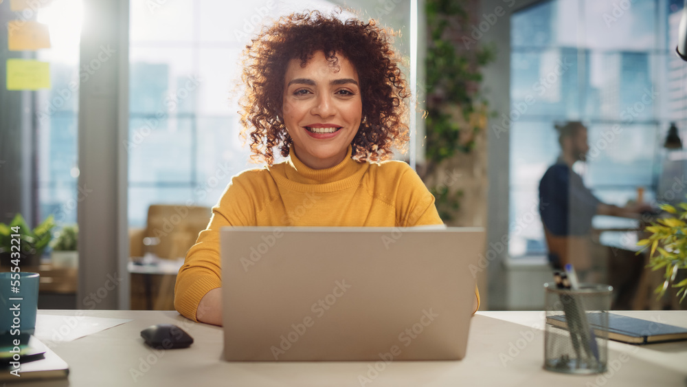Portrait of a Beautiful Middle Eastern Manager Sitting at a Desk in Creative Office. Young Stylish Female with Curly Hair Looking at Camera with Big Smile. Colleagues Working in the Background. - obrazy, fototapety, plakaty 