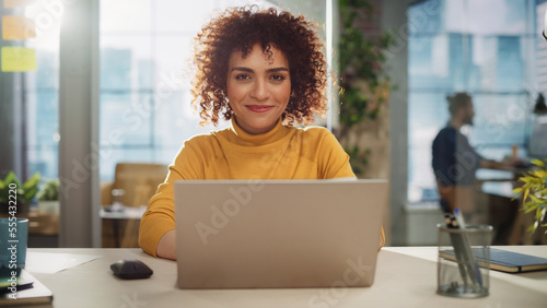 Portrait of a Beautiful Middle Eastern Manager Sitting at a Desk in Creative Office. Young Stylish Female with Curly Hair Looking at Camera with Big Smile. Colleagues Working in the Background. photo