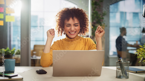 Portrait of a Beautiful Middle Eastern Manager Sitting at a Desk in Creative Office. Young Stylish Female with Curly Hair Celebrating her Achievement with Big Smile