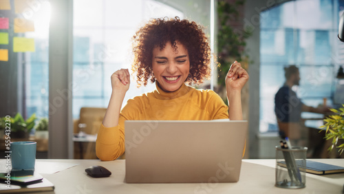 Portrait of a Beautiful Middle Eastern Manager Sitting at a Desk in Creative Office. Young Stylish Female with Curly Hair Celebrating her Achievement with Big Smile photo