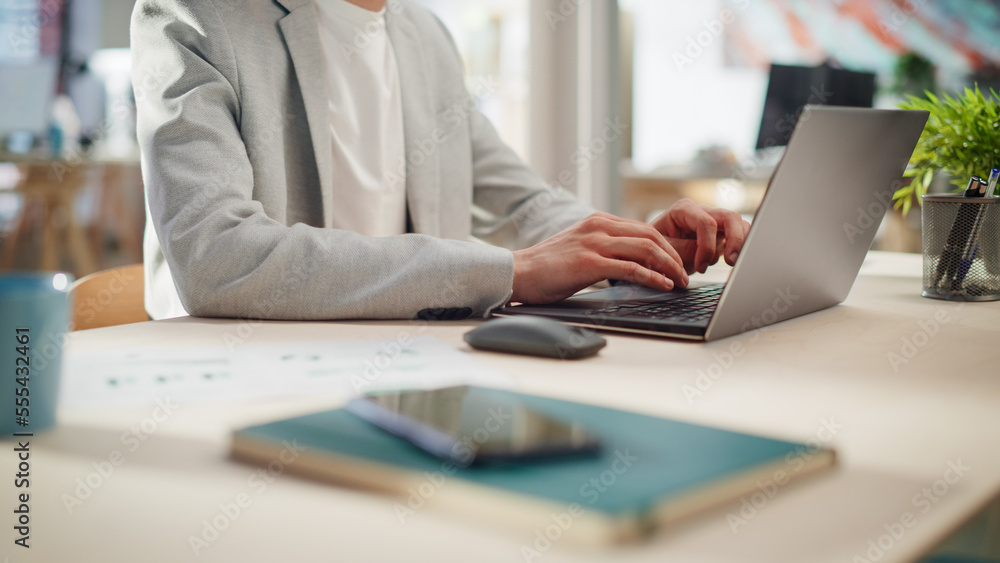 Manager Sitting at a Desk in Creative Office. Focus On Hands of a Young Stylish Man in a Jacket Using Laptop Computer in Marketing Agency. Employee Typing and Using Mouse Manager Sitting at a Desk in Creative Office. Focus On Hands of a Young Stylish Man in a Jacket Using Laptop Computer in Marketing Agency. Employee Typing and Using Mouse