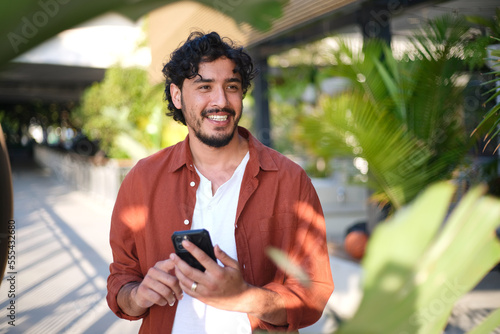 Smiling curly haired man with brown shirt and white tshirt holding his phone