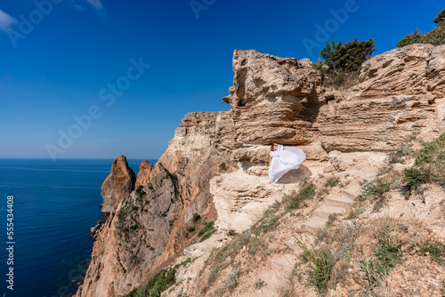 A beautiful young woman in a white light dress with long legs stands on the edge of a cliff above the sea waving a white long dress, against the background of the blue sky and the sea.