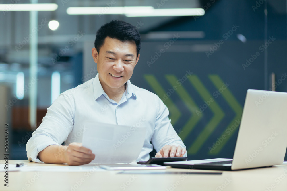 Portrait of a young Asian male accountant sitting at a desk in the office, holding documents in his hands, using a calculator. Works with accounts, budget, loans. Portrait of a young Asian male accountant sitting at a desk in the office, holding documents in his hands, using a calculator. Works with accounts, budget, loans.