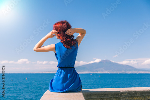 A young girl in a blue dress is sitting on the seashore, in the background the volcano Vesuvius Italy Naples photo