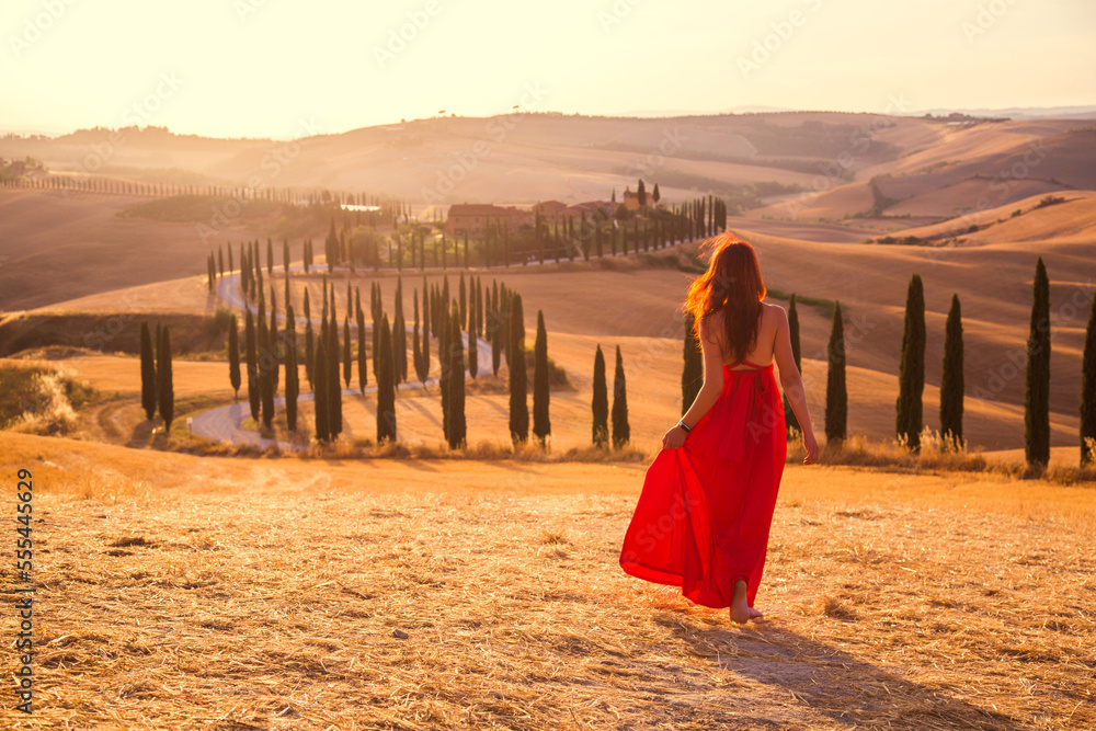 Naklejka premium A girl at sunset in a red dress on a field in Italian Tuscany. Val d'Orcia. Beautiful landscape scenery at sunset of Tuscany in Italy