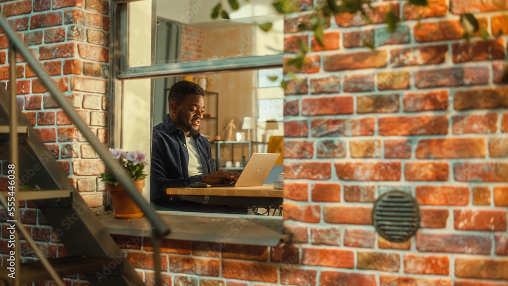 Young Handsome Male Using Laptop Computer, Sitting in Living Room in Apartment. African American Businessman Checking Social Media, Smiling, Answering Emails from Home. Inside Apartment Window View. Young Handsome Male Using Laptop Computer, Sitting in Living Room in Apartment. African American Businessman Checking Social Media, Smiling, Answering Emails from Home. Inside Apartment Window View.