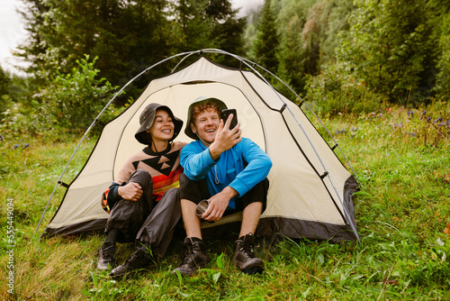Joyful tourist couple taking selfie while sitting at camping tent in green forest