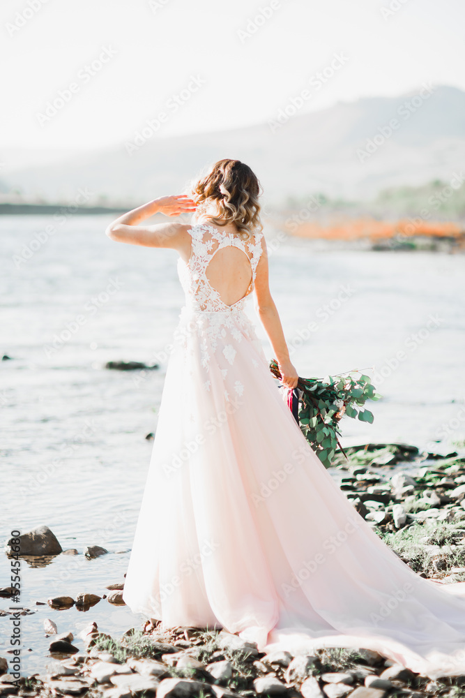 Beautiful brunette bride in elegant white dress holding bouquet posing near river