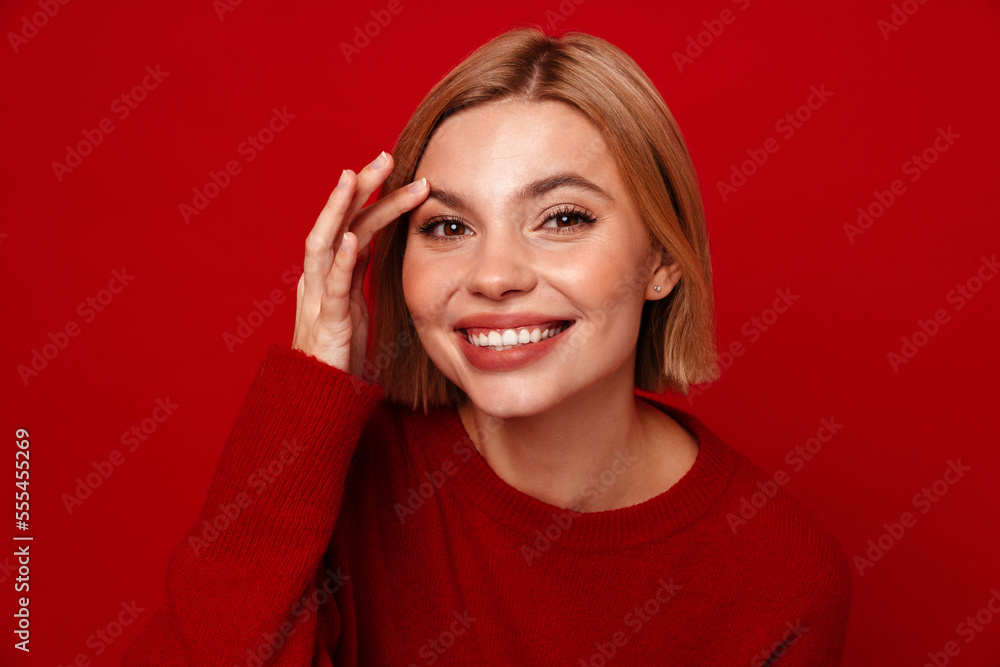 Smiling blonde woman touching her face while standing isolated over red wall Smiling blonde woman touching her face while standing isolated over red wall