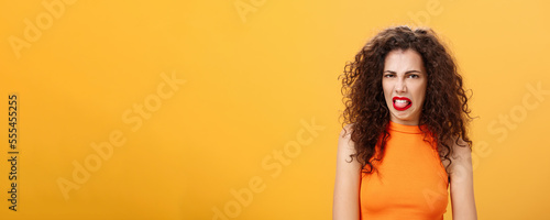 Girl being disgusted with row food sticking out tongue and frowning showing aversion and disgust talking about thing she dislikes with friend standing intense and dissatisfied over orange background