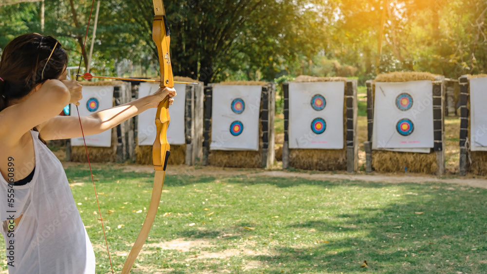 Fototapeta premium Hands of woman aims archery bow and arrow to colorful target in shooting range during training. Exercise and concentration with outdoor archery. Selective focus on hand. Sport and Recreation concept.