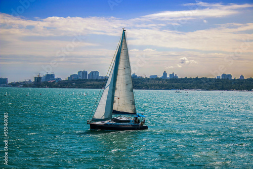 A small sailing yacht sails on the azure sea under a cloudy sky against the backdrop of the city