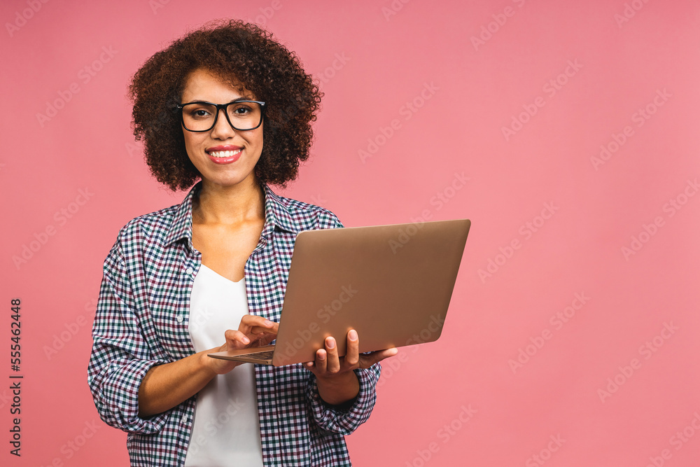 Young african american positive cool lady with curly hair using laptop and smiling isolated over pink background. Young african american positive cool lady with curly hair using laptop and smiling isolated over pink background.