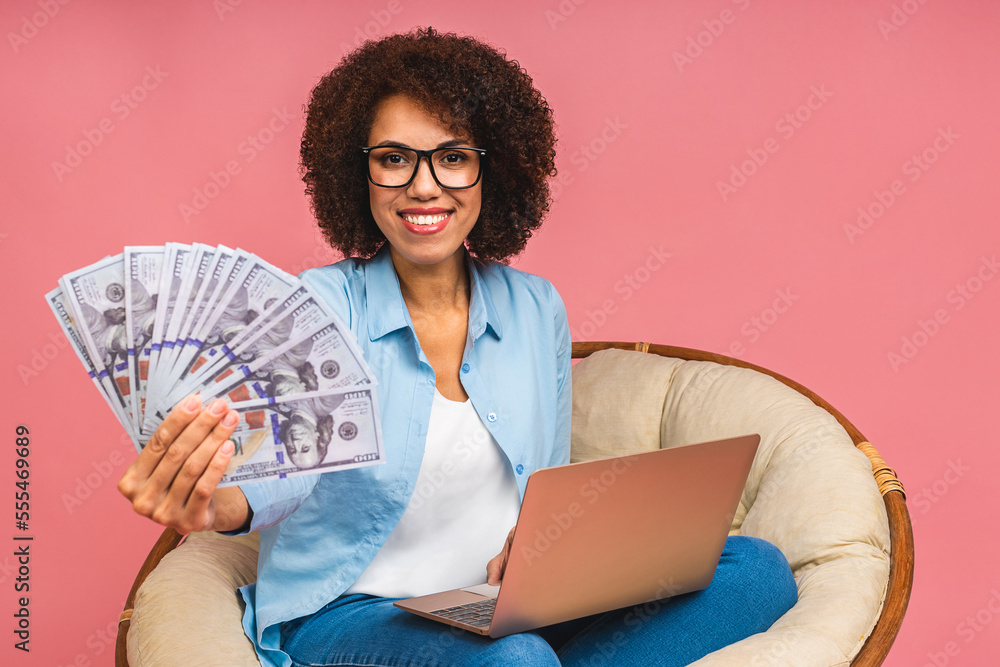 Young african american black positive cool lady with curly hair using laptop and smiling isolated over pink background. Holding money bills. Young african american black positive cool lady with curly hair using laptop and smiling isolated over pink background. Holding money bills.