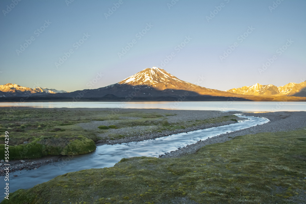 A stream leads the eye towards a high altitude lake. A volcano and snow-capped mountains in the distance are lit by the sunrise against a pure blue sky; Mendoza, Argentina A stream leads the eye towards a high altitude lake. A volcano and snow-capped mountains in the distance are lit by the sunrise against a pure blue sky; Mendoza, Argentina