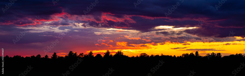 Dramatic sky at sunset with dark clouds and colourful light glowing above silhouetted trees; Bromont, Quebec, Canada Dramatic sky at sunset with dark clouds and colourful light glowing above silhouetted trees; Bromont, Quebec, Canada