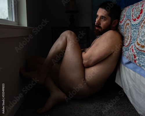 Fotografie attractive male model sitting naked on floor against bed holding himself with mu