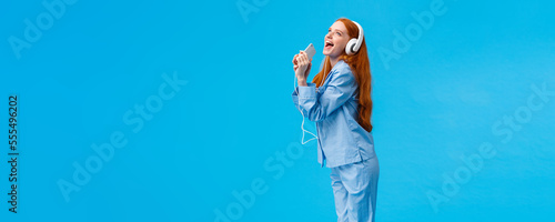 Girl dreaming become star. Vertical full-length portrait charming and carefree redhead teenager in nightwear, donwload karaoke app, listening music in headphones, singing into smartphone photo