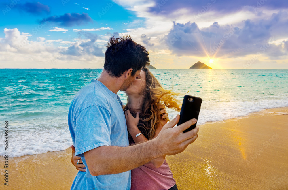 A couple stands kissing on Lanakai beach on the Hawaiian island of Oahu while holding a smart phone and taking a self-portrait at sunset; Lanakai, Oahu, Hawaii, United States of America A couple stands kissing on Lanakai beach on the Hawaiian island of Oahu while holding a smart phone and taking a self-portrait at sunset; Lanakai, Oahu, Hawaii, United States of America