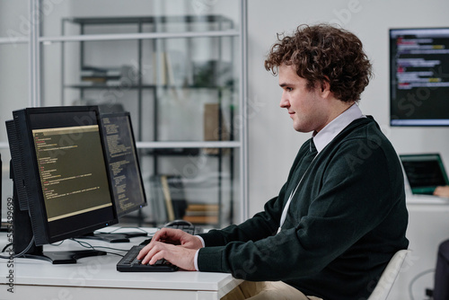 Young IT specialist writing codes for program on computer while sitting at his workplace in IT office