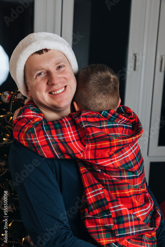 Dad and son in red pajamas near the Christmas tree and hugging. Front view © Olha