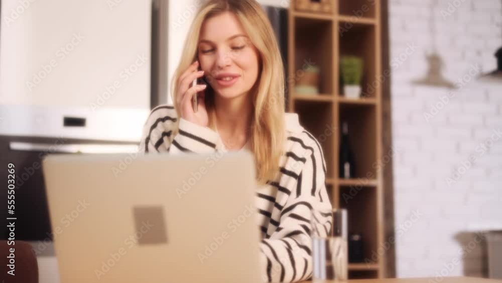 Dreamy soft shot of busy smiling freelancer self employee woman talking by smartphone mobile phone having working or personal conversation sitting at home workplace at laptop computer desk indoors Dreamy soft shot of busy smiling freelancer self employee woman talking by smartphone mobile phone having working or personal conversation sitting at home workplace at laptop computer desk indoors