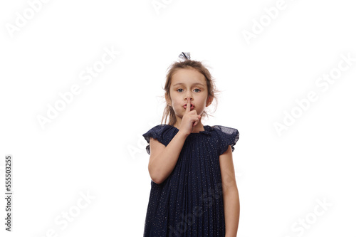 Adorable Caucasian baby girl in elegant navy dress, showing CHUT on camera, putting her finger on her lips, looking confidently at camera, isolated over white background with free advertising space