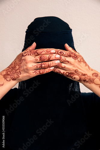 Close-up portrait of woman wearing black muslim hijab and muslim dress, hands covering eyes and showing hands painted with henna in arabic style, studio shot on whtie background photo