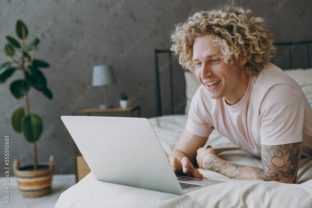Young happy fun IT man wear white t-shirt pajama he lying in bed hold use work on laptop pc computer study online rest relax spend time in bedroom home in own room hotel wake up. Real estate concept.
