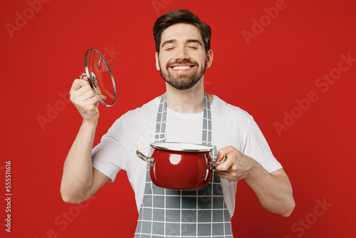 Young fun male housewife housekeeper chef cook baker man wear grey apron hold in hand pot lid open saucepan sniff scent enjoy close eyes isolated on plain red background studio. Cooking food concept. photo