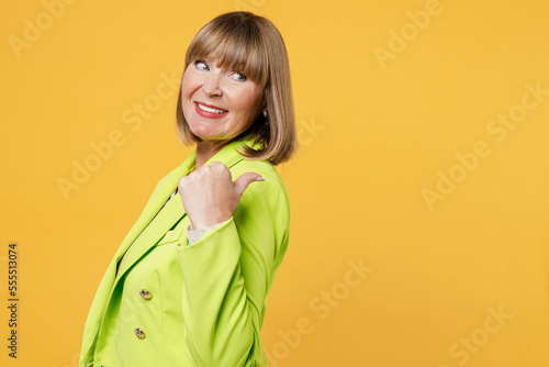 Side view elderly happy woman 50s years old wear green jacket white t-shirt point thumb finger aside indicate on workspace area copy space mock up isolated on plain yellow background studio portrait.