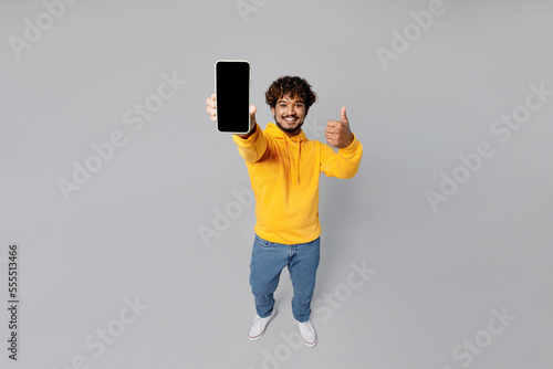 Obraz na plátne Full body smiling overjoyed happy fun cheerful young Indian man 20s he wearing casual yellow hoody hold in hand use mobile cell phone show thumb up isolated on plain grey background studio portrait
