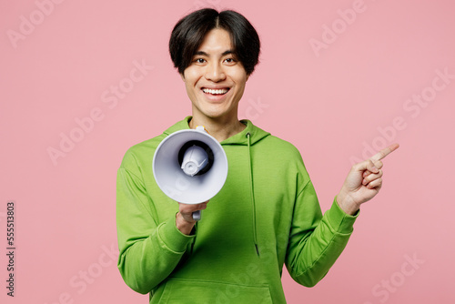 Young happy fun man of Asian ethnicity wear green hoody hold in hand megaphone scream announces discounts sale Hurry up point index finger aside on area isolated on plain pastel light pink background.