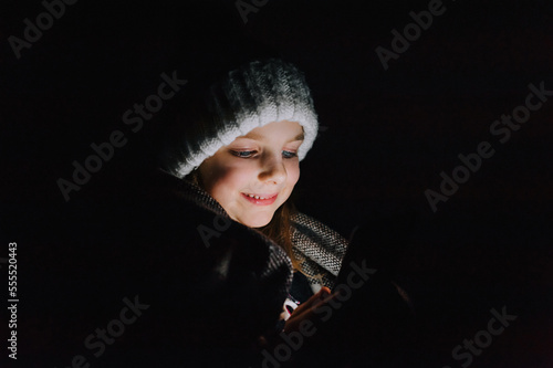 Blackout in Ukraine. Ukrainian beautiful girl, a child in warm clothes, a hat, a blanket, sits in a dark, cold room, looks at a smartphone, phone and plays games. Photography, portrait.