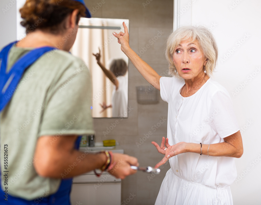 Fototapeta premium Stressed elderly woman talking with plumber and gesturing emotionally in her home