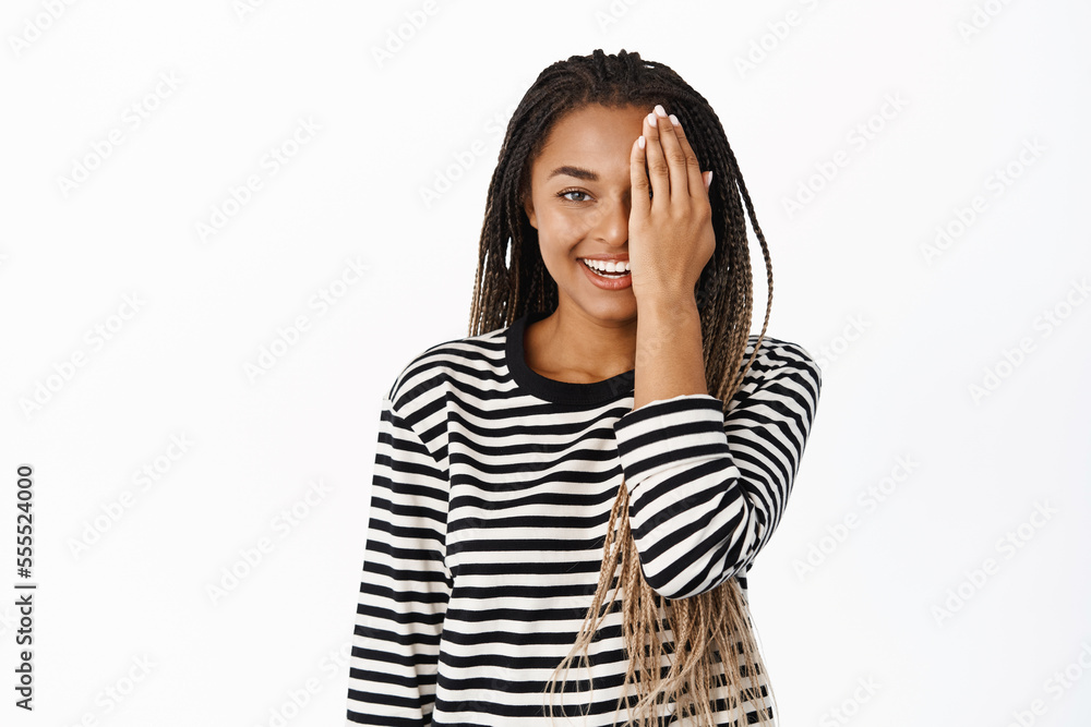 Smiling black girl covers half of her face behind palm and smiles at camera, wears casual clothes, white background. Before after concept Smiling black girl covers half of her face behind palm and smiles at camera, wears casual clothes, white background. Before after concept