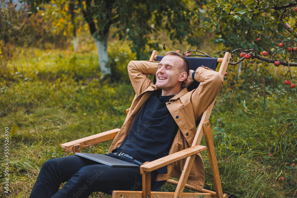 A young man works on a laptop outdoors. A young freelancer rests in the forest. Remote work, active recreation in the summer. Tourism, people concept - man sitting on a chair outdoors. A young man works on a laptop outdoors. A young freelancer rests in the forest. Remote work, active recreation in the summer. Tourism, people concept - man sitting on a chair outdoors.