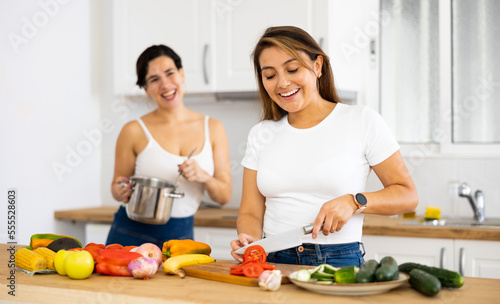 Cheerful young Hispanic woman with female roommate cooking together in home kitchen, preparing vegetable dish for dinner